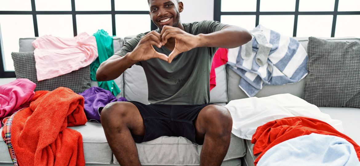 guy holding his hands up in a heart shape while sitting on a living room couch with laundry covering the cushions