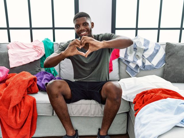 guy holding his hands up in a heart shape while sitting on a living room couch with laundry covering the cushions